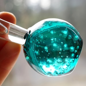 May include: A teal glass bottle stopper with a round, textured top. The stopper has a clear glass tube attached to the bottom.