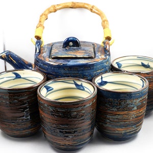 Op de afbeelding: Een blauwe en bruine theepot van keramiek met een bamboe handvat en vier bijpassende theekopjes. De kopjes hebben een witte binnenkant met blauwe en witte strepen en een blauw vogeltje ontwerp.