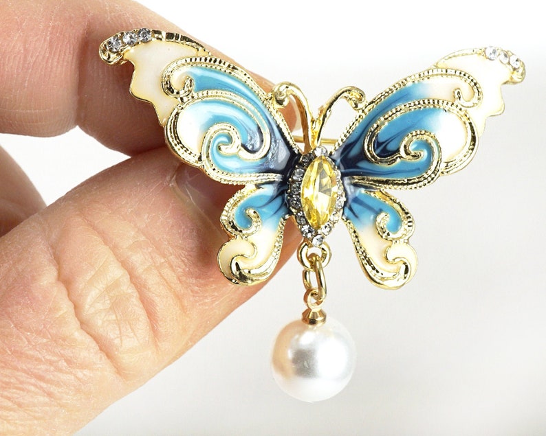 May include: A gold-tone butterfly brooch with blue and white enamel and a dangling pearl. The butterfly has intricate details and a clear rhinestone in the centre.