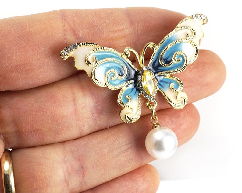 May include: A gold-tone butterfly brooch with blue and white enamel and a dangling pearl. The butterfly has intricate details and a clear rhinestone in the centre.