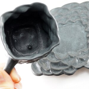 May include: A gray ceramic teacup and saucer set with a textured, floral design. The cup has a handle and a small, round indentation on the bottom.