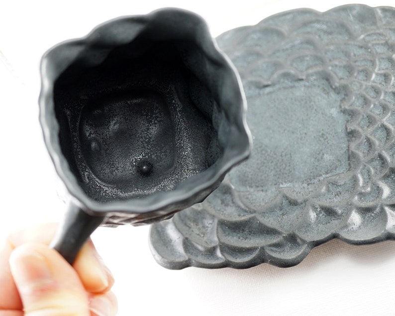 May include: A gray ceramic teacup and saucer set with a textured, floral design. The cup has a handle and a small, round indentation on the bottom.