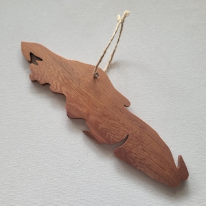 May include: A wooden ornament shaped like the outline of an island. The ornament is brown and has a natural wood grain. It has a hole at the top for hanging.