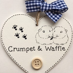 May include: White wooden heart-shaped sign with a blue and white gingham bow. The sign has a black and white illustration of two guinea pigs, paw prints, and the text "Crumpet & Waffle".