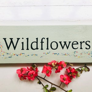 May include: A light green wooden sign with the word "Wildflowers" painted in black. The sign has a painted border of small colourful flowers.