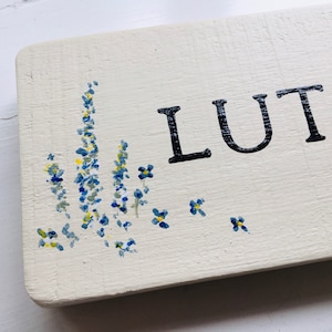 May include: A white wooden sign with the word "LUTO" painted in black. The sign is decorated with blue and yellow flowers painted on the bottom.