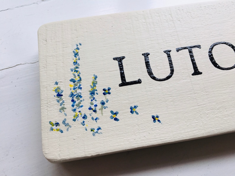May include: A white wooden sign with the word "LUTO" painted in black. The sign is decorated with blue and yellow flowers painted on the bottom.