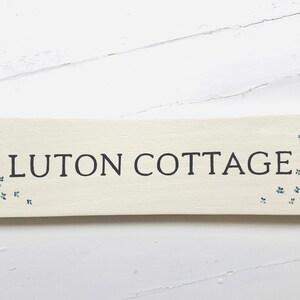 May include: A white wooden sign with the text "LUTON COTTAGE" in black lettering. The sign is decorated with small blue and yellow flowers.