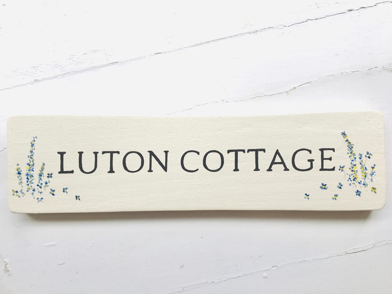 May include: A white wooden sign with the text "LUTON COTTAGE" in black lettering. The sign is decorated with small blue and yellow flowers.