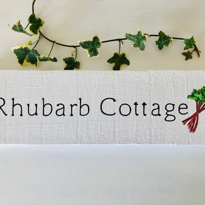 May include: White wooden sign with the text "Rhubarb Cottage" painted in black. A small painted image of rhubarb stalks with green leaves is on the right side of the sign.