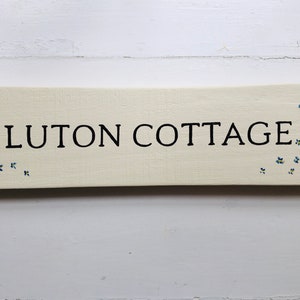 May include: A white wooden sign with black lettering that reads "Luton Cottage". The sign is decorated with blue and yellow flowers.
