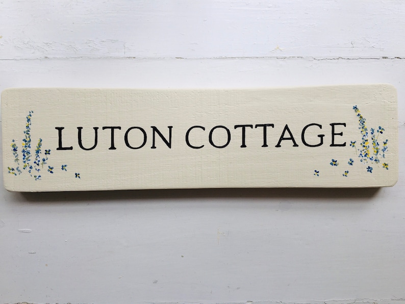 May include: A white wooden sign with black lettering that reads "Luton Cottage". The sign is decorated with blue and yellow flowers.