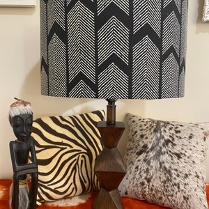 May include: A black and white lampshade with a chevron pattern sits on a wooden base. The lamp is on a table with a zebra print pillow and a cowhide pillow.
