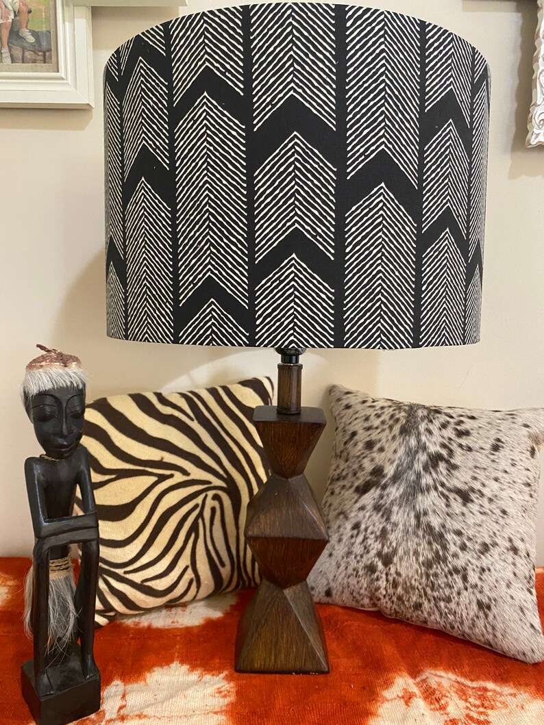May include: A black and white lampshade with a chevron pattern sits on a wooden base. The lamp is on a table with a zebra print pillow and a cowhide pillow.