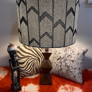 May include: A black and white lampshade with an arrow pattern sits on a wooden base. The lamp is on a table with a red and white patterned surface. There are two pillows on the table, one with a zebra print and the other with a cowhide pattern. A wooden figurine of a person is on the left side of the table.