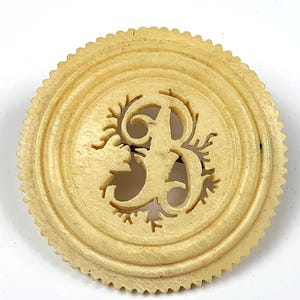 May include: A circular, cream-coloured vintage brooch featuring an openwork letter "B" at its centre. The brooch has a ridged border and a scalloped edge. The design is detailed and intricate.