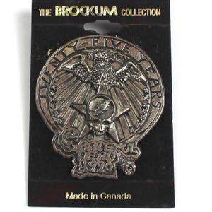 May include: A silver-colored metal pin with an eagle, skull, and star design. The pin is on a black card with the text "THE BROCKUM COLLECTION" and "TWENTY FIVE YEARS". The text "GRATEFUL DEAD 1970" is also visible.