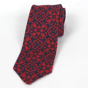 Może przedstawiać: Zwinięty krawat z powtarzającym się geometrycznym wzorem w kolorze czerwonym i granatowym. Krawat wykonany jest z teksturowanej tkaniny i leży na białej powierzchni. Klasyczny dodatek.
