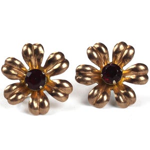 Op de afbeelding: Een paar goudkleurige bloemvormige oorstekers. Elke oorbel heeft zes bloemblaadjes en een donkerrode edelsteen in het midden. De oorbellen staan tegen een witte achtergrond.