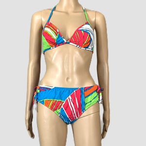 May include: A two-piece swimsuit with a vibrant abstract design. The halter-neck bikini top features a mix of red, green, blue, and white patterns. The matching bottoms have tie sides and a similar design. The swimsuit is displayed on a mannequin.
