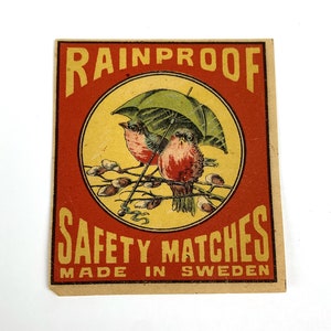 May include: Vintage matchbox label with a red background and the text "RAINPROOF SAFETY MATCHES MADE IN SWEDEN". The label features a yellow circle with two birds perched on a branch under a green umbrella.