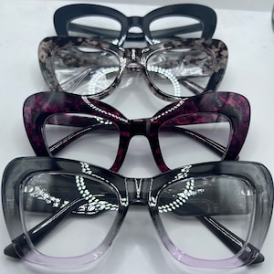May include: Four pairs of cat-eye glasses in different colours and patterns. The glasses are stacked on top of each other, with the bottom pair being the most visible. The glasses are all black, with the exception of the second pair, which is a light grey with a black pattern.