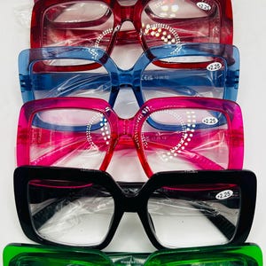 Puede incluir: Una pila de cinco pares de gafas de lectura con monturas cuadradas. Las monturas son de diferentes colores, incluyendo rojo, azul, rosa, negro y verde. Cada par tiene una pegatina en la lente derecha que dice +2,25.