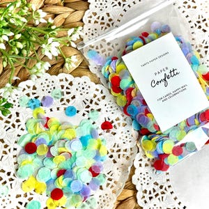May include: A clear plastic bag filled with colorful paper confetti. The confetti is in various colors, including red, yellow, blue, and green. A white card with the text "Tarts Paper Designs Paper Confetti" is inside the bag. The confetti is scattered on a white lace doily.