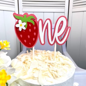 May include: A pink and white cake topper with the word "one" and a red strawberry with a white flower. The topper is on a white cake with white coconut flakes.