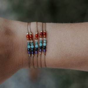 May include: A set of seven bracelets with different colored beads, including red, orange, yellow, green, blue, purple, and clear. The bracelets are made with thin brown string and are stacked on a person's wrist.
