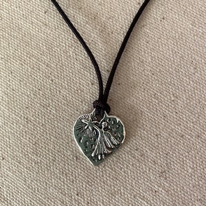 May include: A silver-colored pendant necklace featuring a heart-shaped charm with a raised design of a figure holding a star. The charm is suspended from a black cord.