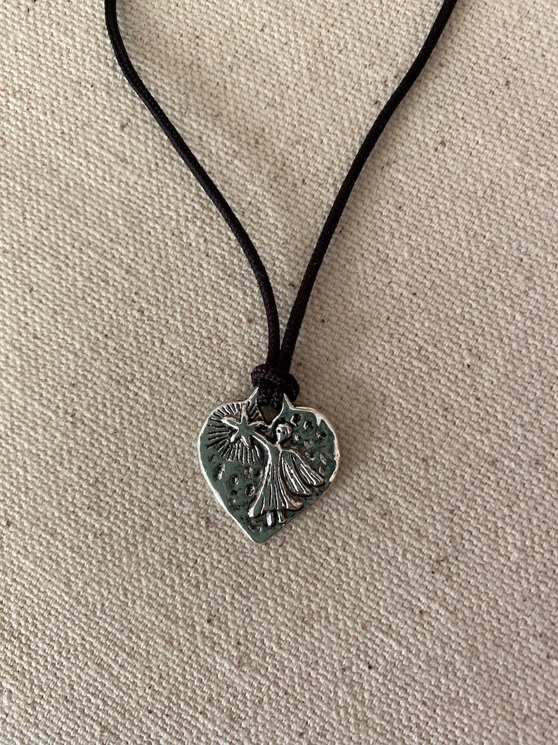 May include: A silver-colored pendant necklace featuring a heart-shaped charm with a raised design of a figure holding a star. The charm is suspended from a black cord.