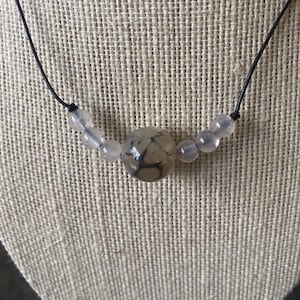 May include: A necklace featuring a large, round, patterned bead as the focal point, flanked by smaller, translucent beads. The beads are strung on a black cord, creating a simple yet elegant design. The central bead has a unique, marbled appearance.