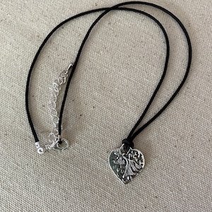 May include: A silver-colored heart pendant necklace with a black cord. The heart pendant features a detailed design of two figures and a star. The necklace has a silver-colored chain for adjusting the length.