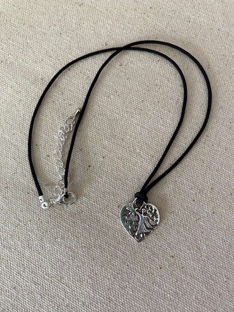 May include: A silver-colored heart pendant necklace with a black cord. The heart pendant features a detailed design of two figures and a star. The necklace has a silver-colored chain for adjusting the length.