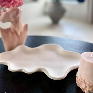 Puede incluir: Arreglo decorativo con un jarr&oacute;n de cer&aacute;mica rosa en forma de busto, con flores rosas y blancas. Tambi&eacute;n hay una bandeja de cer&aacute;mica blanca en forma de nube y una vela rosa con detalles florales.