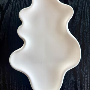 Puede incluir: Un plato de cer&aacute;mica blanco con forma org&aacute;nica. El plato tiene una superficie lisa y un dise&ntilde;o fluido con bordes redondeados. Se encuentra sobre un fondo de vetas de madera oscura, creando un contraste de texturas.
