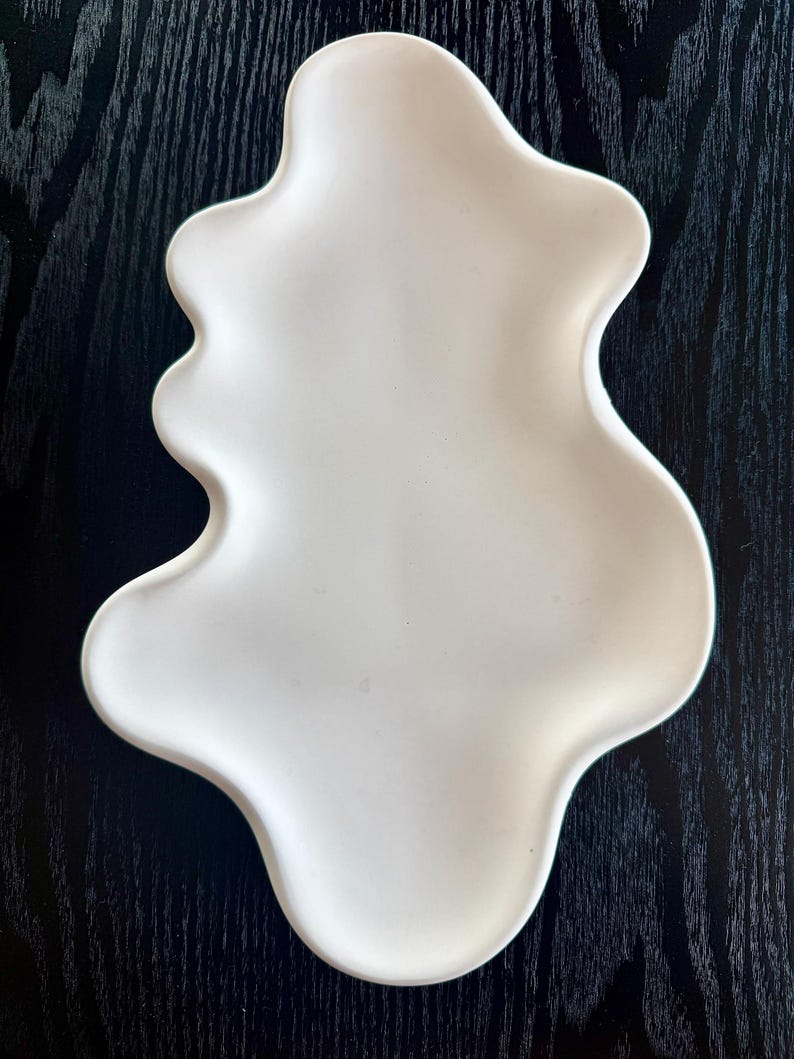 Puede incluir: Un plato de cer&aacute;mica blanco con forma org&aacute;nica. El plato tiene una superficie lisa y un dise&ntilde;o fluido con bordes redondeados. Se encuentra sobre un fondo de vetas de madera oscura, creando un contraste de texturas.