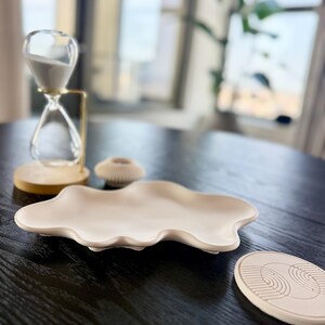 Puede incluir: Una disposici&oacute;n decorativa sobre una mesa de madera oscura. Incluye un reloj de arena transparente con arena blanca, una fuente de cer&aacute;mica beige con forma de nube, un peque&ntilde;o portavelas beige y un posavasos beige con dise&ntilde;o geom&eacute;trico.