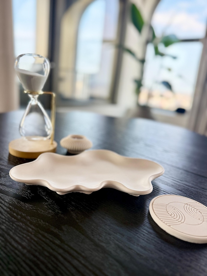 Puede incluir: Una disposici&oacute;n decorativa sobre una mesa de madera oscura. Incluye un reloj de arena transparente con arena blanca, una fuente de cer&aacute;mica beige con forma de nube, un peque&ntilde;o portavelas beige y un posavasos beige con dise&ntilde;o geom&eacute;trico.