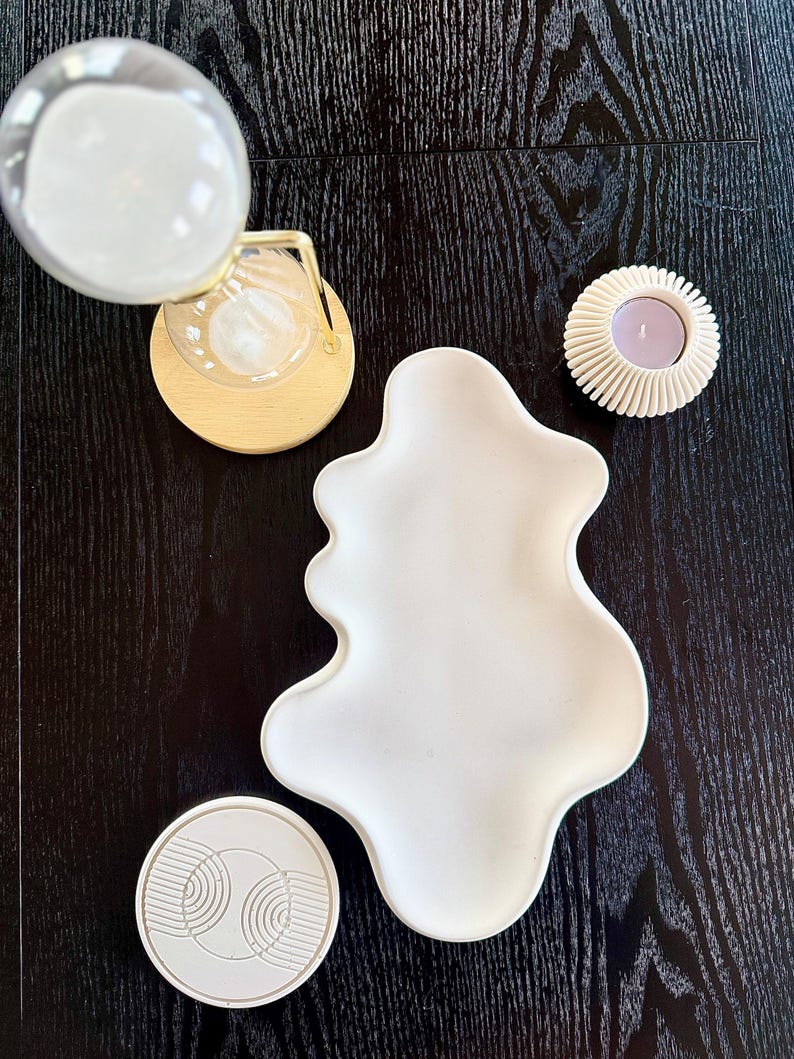 Puede incluir: Una variedad de art&iacute;culos de decoraci&oacute;n del hogar sobre una superficie de madera oscura. Incluye un reloj de arena transparente con un marco de metal dorado, un plato blanco de forma org&aacute;nica, un posavasos blanco con un dise&ntilde;o geom&eacute;trico y un portavelas blanco con una vela morada.