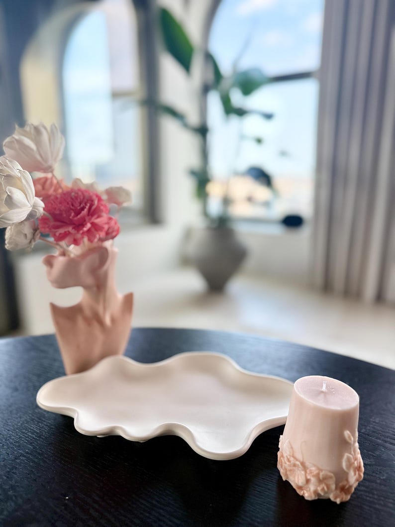 Puede incluir: Arreglo decorativo con un jarr&oacute;n de cer&aacute;mica rosa en forma de busto, con flores rosas y blancas. Tambi&eacute;n hay una bandeja de cer&aacute;mica blanca en forma de nube y una vela rosa con detalles florales.