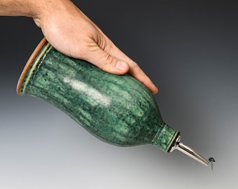 May include: A ceramic oil cruet with a mottled green glaze and a smooth, rounded body. The vessel features a narrow neck topped with a silver-colored metal pour spout, designed for controlled dispensing of liquids from the tapered opening.