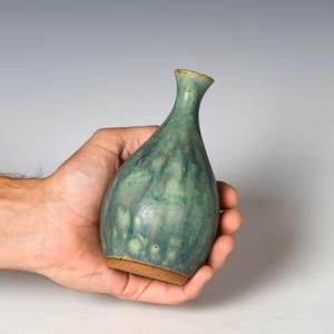 May include: A small ceramic bud vase with a narrow neck and a bulbous body. The surface features a mottled glaze in shades of teal, sage green, and hints of brown, with an unglazed, textured terracotta-colored base at the bottom.