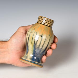 May include: A small ceramic vase featuring a flared base and a narrow neck with horizontal black line detailing. The body displays a blended glaze of earthy tan, muted green, and deep blue tones that drip vertically down the surface.