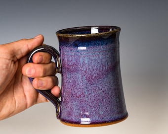 May include: A ceramic mug featuring a speckled glaze with a blend of deep purple, light blue, and magenta tones. The mug has a cylindrical body, a wide handle, and a dark, glossy rim that transitions into the mottled purple and blue body color.