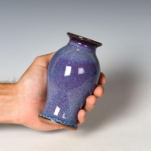 May include: A small ceramic vase with a flared rim and a rounded body, featuring a mottled glaze in shades of deep purple, violet, and blue with a smooth, glossy finish and a visible brown clay base at the bottom edge.