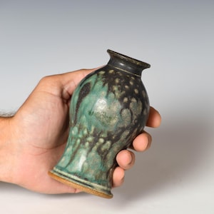 May include: A small ceramic vase with a flared rim and a tapered neck. The surface features a mottled glaze with deep black and forest green tones, creating a textured, layered appearance. The base of the vase reveals a raw, unglazed brown clay.
