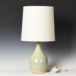 Può includere: Una lampada da tavolo con base in ceramica a forma di goccia con una finitura lucida maculata verde chiaro e blu. La lampada è completata da un paralume cilindrico alto in tessuto bianco e include un cavo elettrico bianco con una spina.