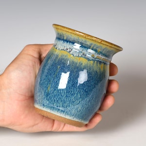 May include: A small ceramic cup with a flared rim, featuring a glossy glaze in shades of deep blue, teal, and golden yellow. The surface has a textured, mottled appearance with visible drips and a smooth, unglazed brown clay base.